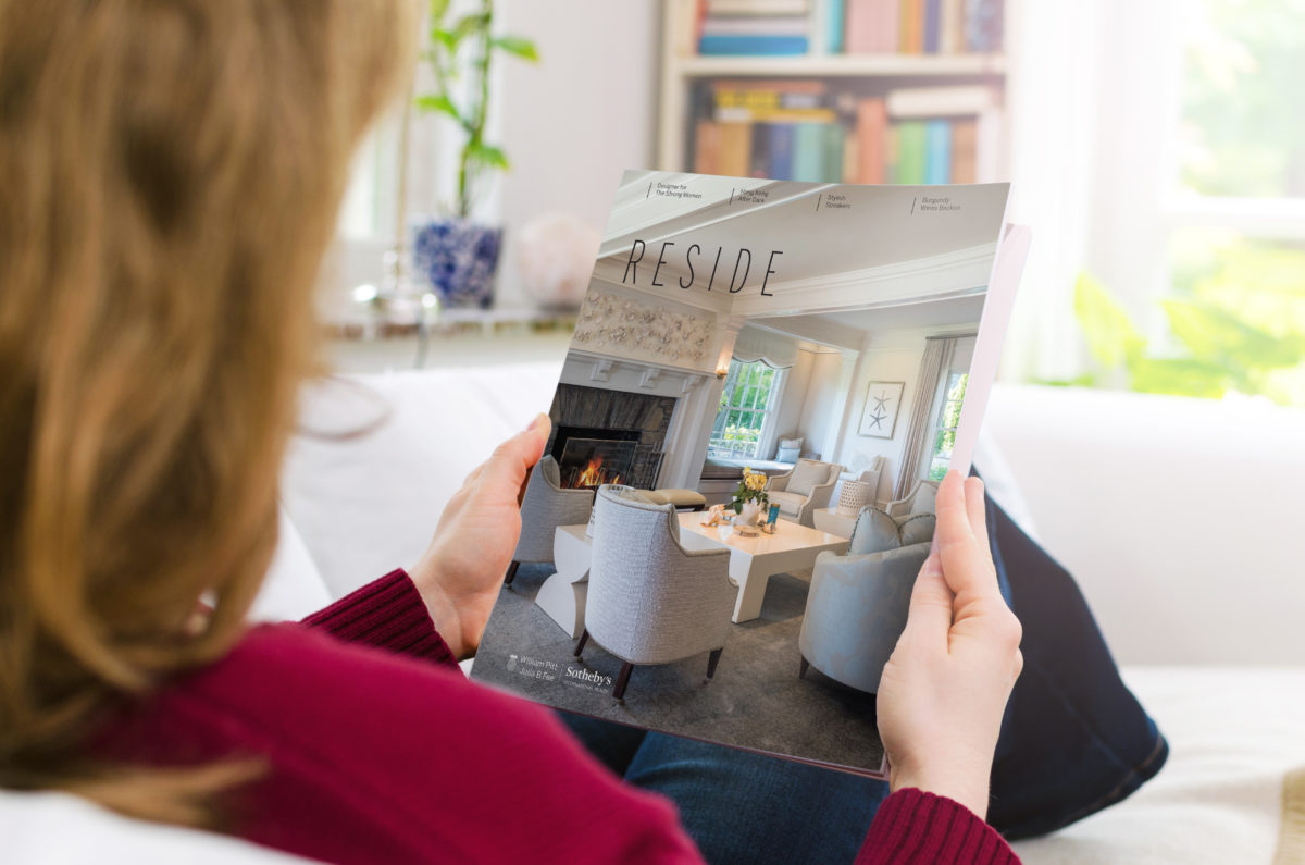 Blond woman holding a blank brochure or book cover with copy space. The photo was taken indoors in a domestic room by photographing over the woman's shoulder.